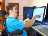 Spastic young man with infantile cerebral palsy caused by a complicated birth sitting in a multifunctional wheelchair, using a computer with a wireless headset, reaching out to touch the touch screen.